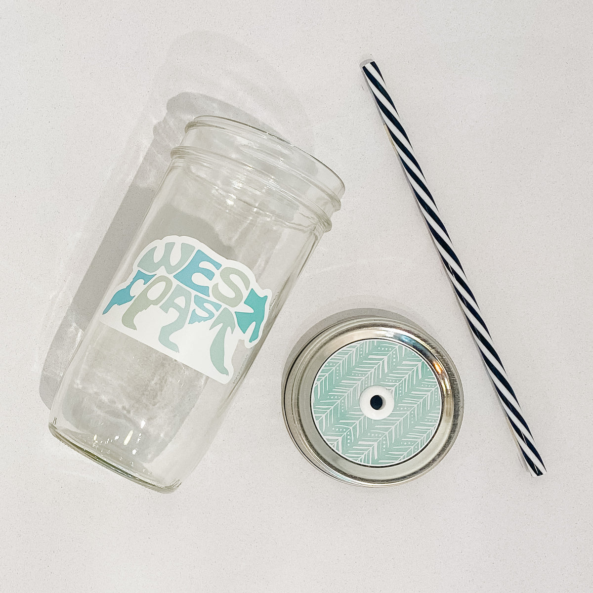 Flat lay of mason jar tumbler that reads west coast in shape of bear, colour is multiple shades of blues. Black and white striped straw. Mason jar straw lid with white boho arrows with a aqua background.