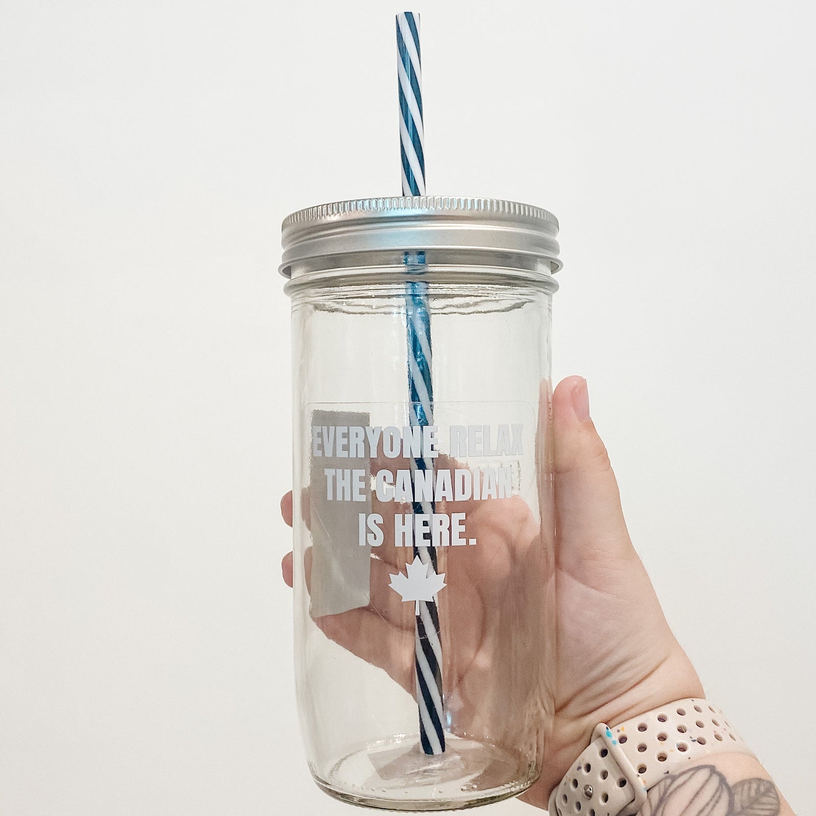 Hand holding mason jar that reads everyone relax the Canadian is here with white text with small maple leaf at bottom.