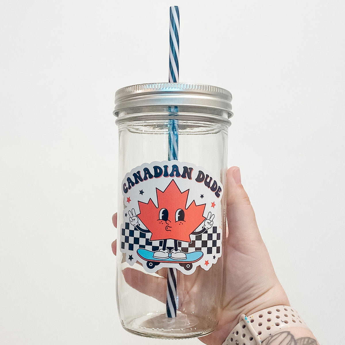 Hand holding mason jar that reads Canadian dude in a retro font with an animated maple leaf riding a small blue skateboard.