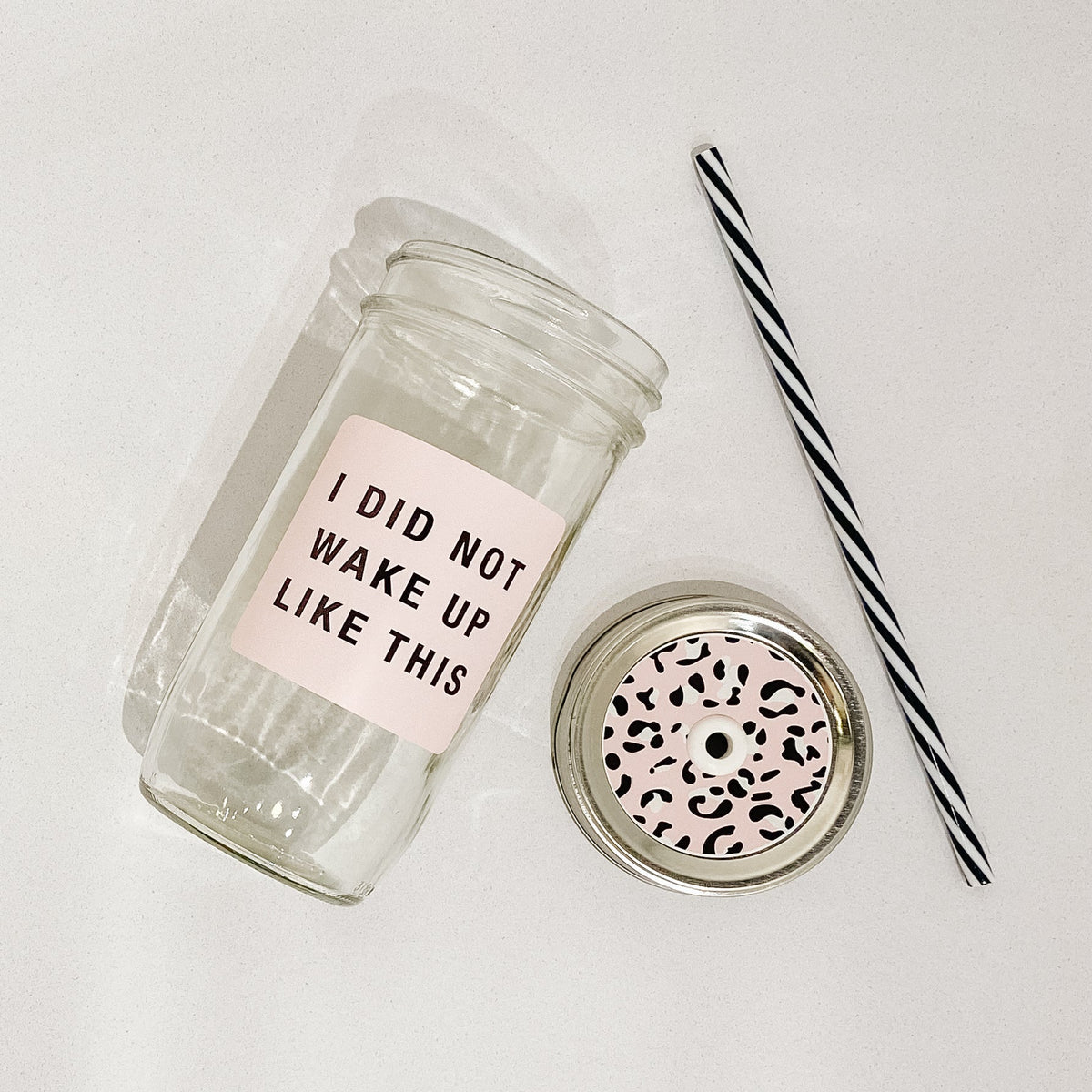 Flat lay of a mason jar tumbler that says "I did not wake up like this” in black on a pink background, a pink cheetah print straw lid, and a black and white striped straw