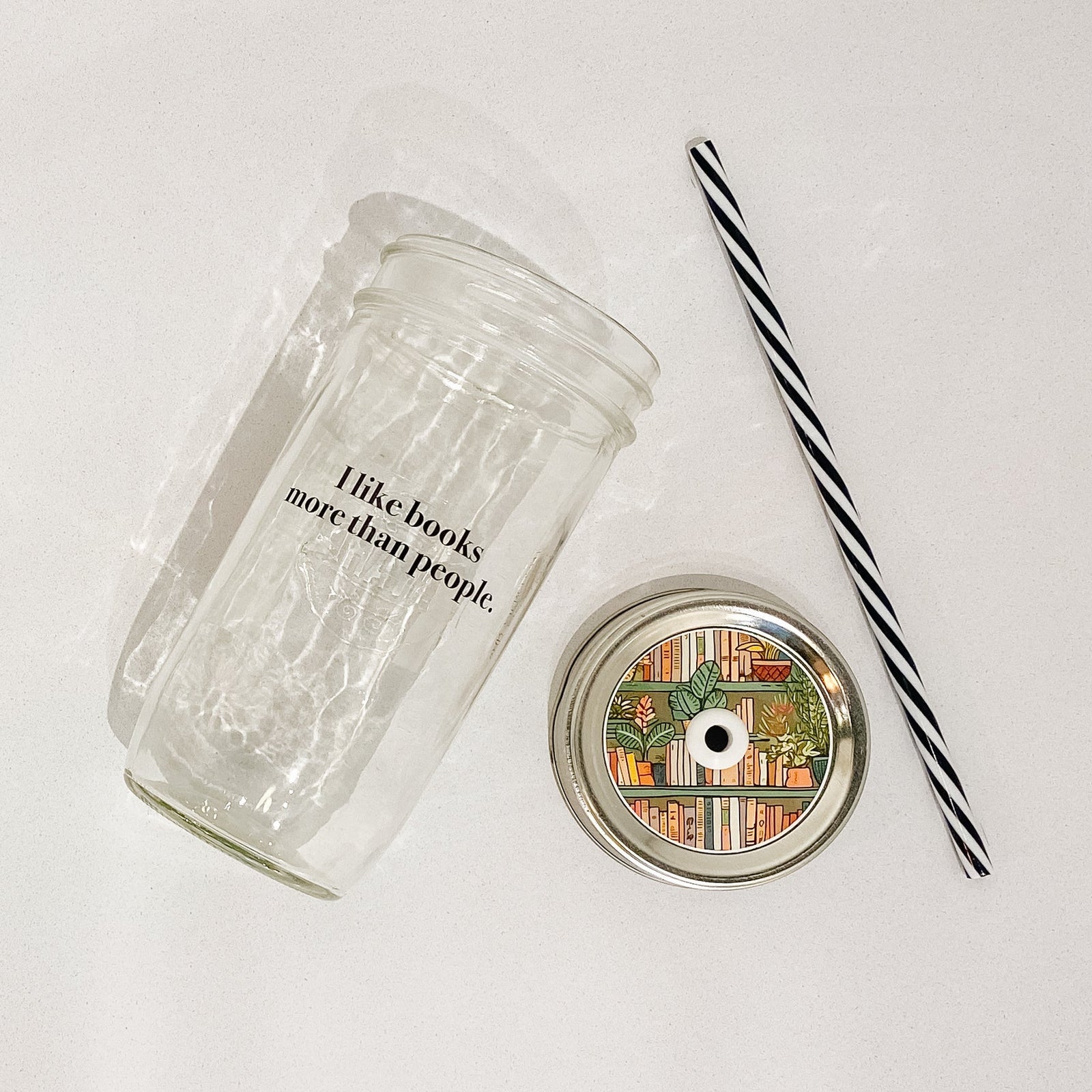 Flat lay of a mason jar tumbler that says "I like books more than people”, a straw lid this a bookshelf scene on it, and a black and white striped straw.