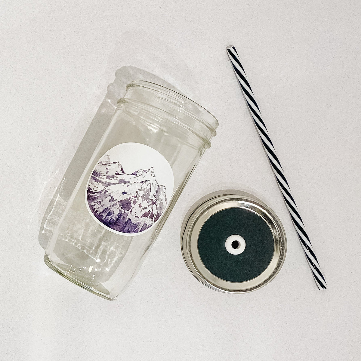 Flat lay of a mason jar tumbler that has a circle decal of a snowy mountain peak, a straw lid that is a charcoal grey, and a black and white striped straw.