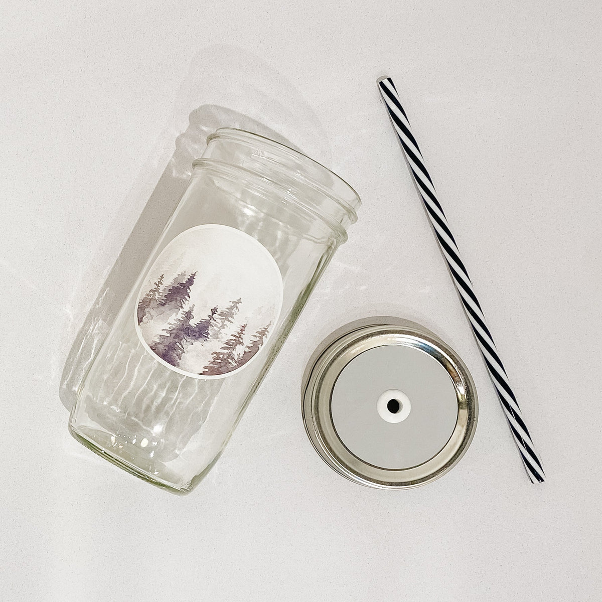 Flat lay of a mason jar tumbler that has a circle decal of trees in fog, a straw lid that is a very pale grey, and a black and white striped straw