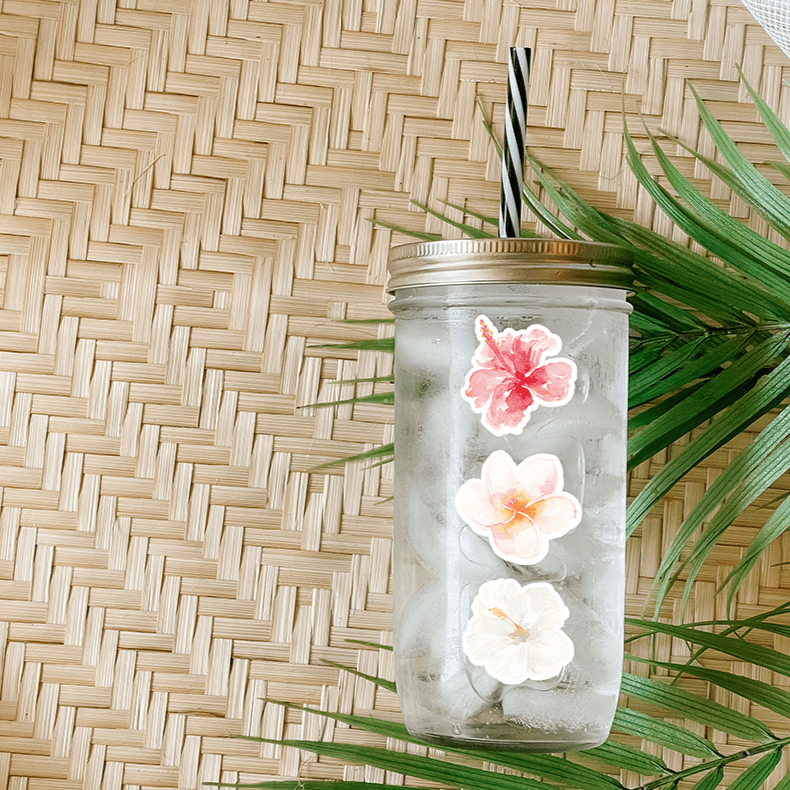 Tumbler with water and a sticker that has a pink, peach, and white hibiscus flowers. Photographed as a flat lay in a winnowing tray with palm leaves.