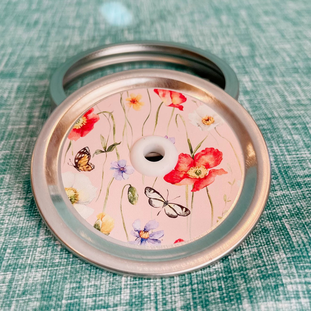 straw lid with a pink background, there are green stems/leaves taking up most of the centre of the lid, there are also flowers and butterflies scattered