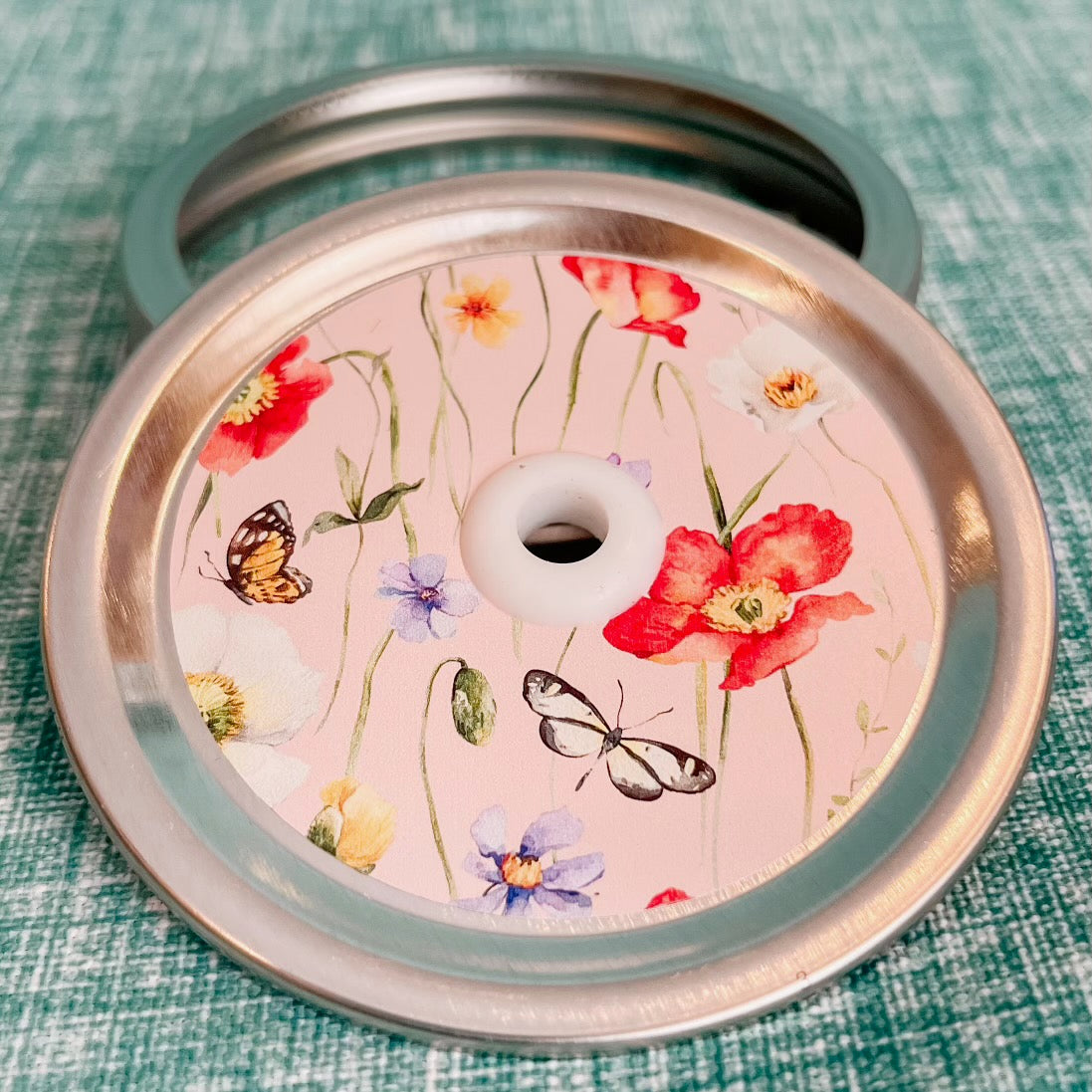 close up of a straw lid with a pink background, there are green stems/leaves taking up most of the centre of the lid, there are also flowers and butterflies scattered