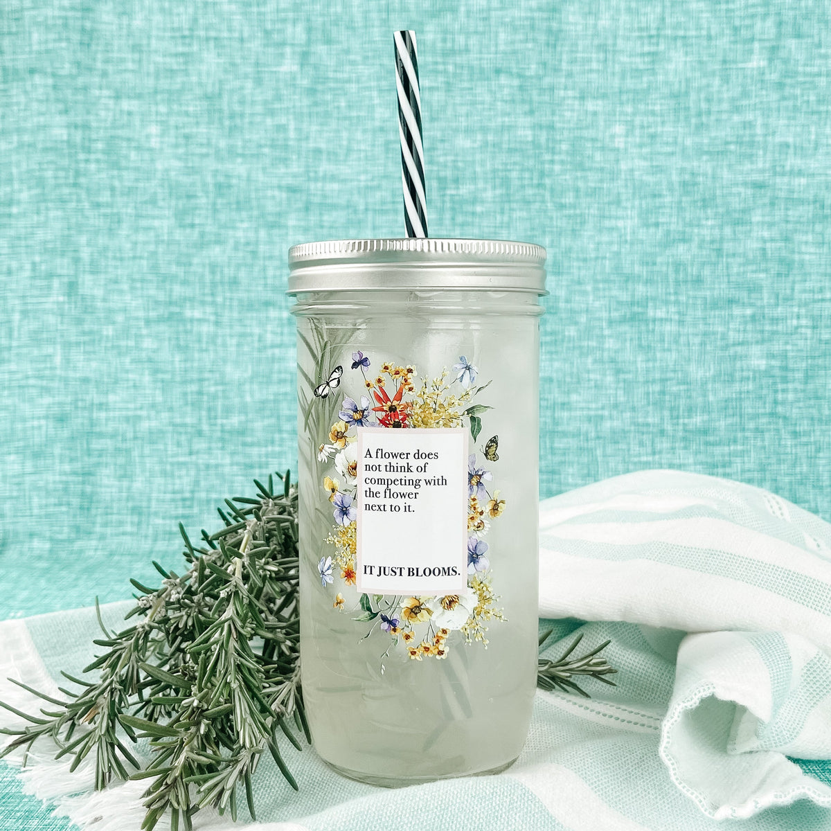 Mason jar tumbler that says “A flower does not think of competing with the flower next to it. It just blooms.” With flowers creating a rectangle around the words.