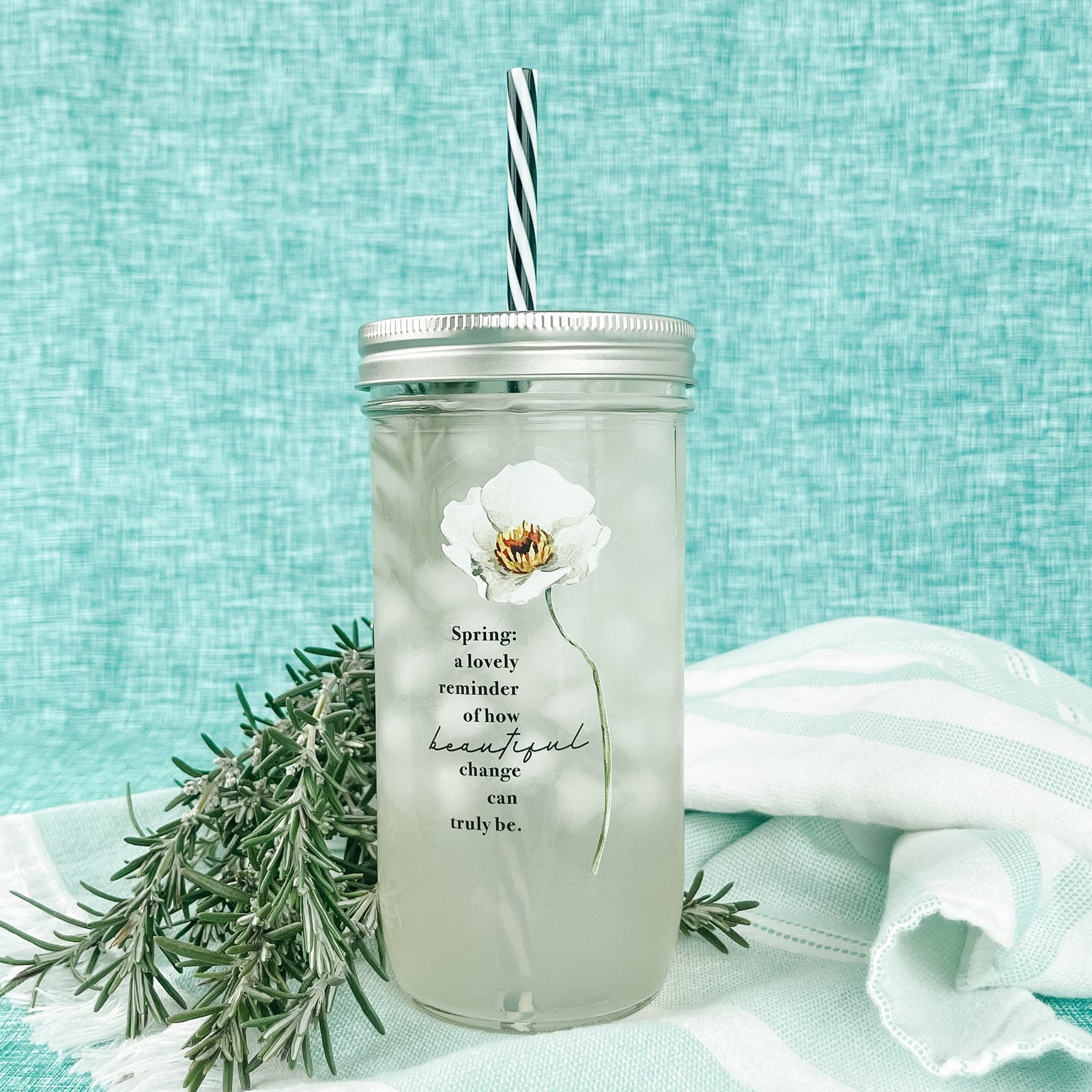 Mason jar tumbler that says “Spring: a lovely reminder of how beautiful change can truly be.” With a large white flower to the right of the words