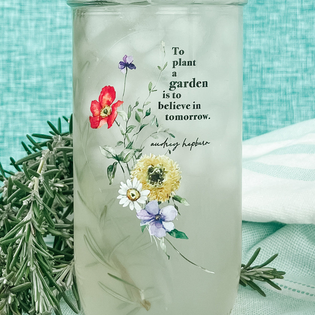 Close up of a mason jar tumbler that says “To plant a garden is to believe in tomorrow. Audrey Hepburn” with florals to the bottom left of the quote.