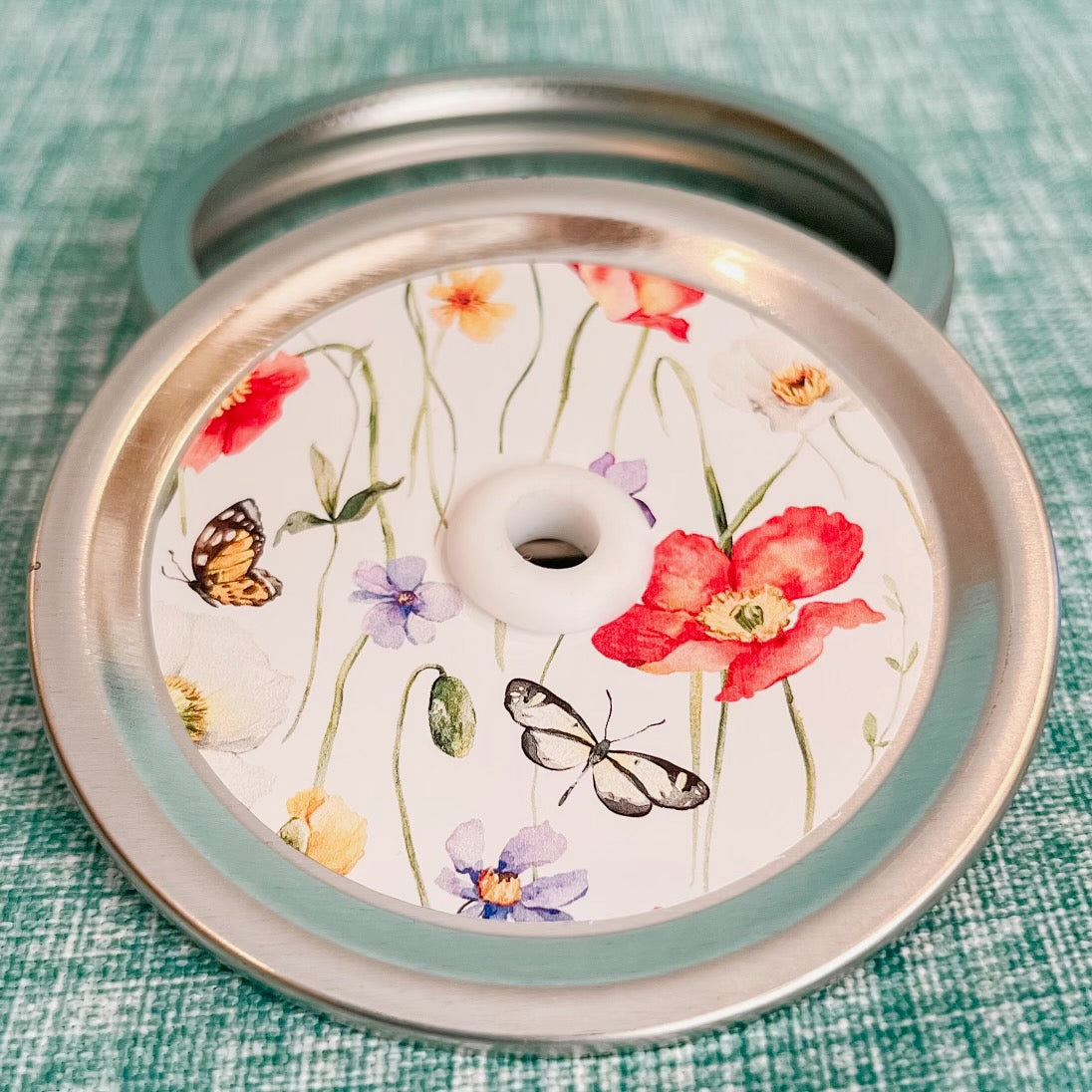straw lid with a white background, there are green stems/leaves taking up most of the centre of the lid, there are also flowers and butterflies scattered
