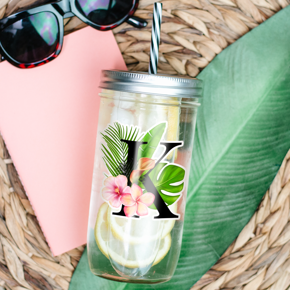 Transparent Tumbler with water and lemon inside and a sticker of flower, coconut leaf and ornamental plants with big letter K printed on it. Tumbler is photographed laying on a table with banana leaf and sunglasses beside it.