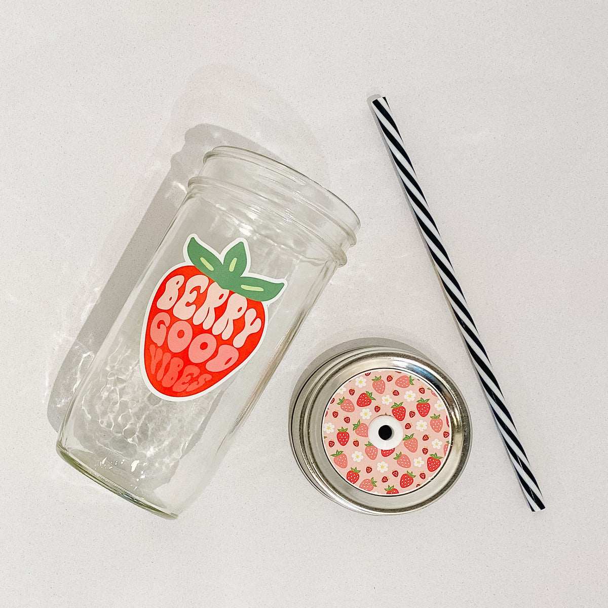 Flat lay of a mason jar tumbler that says “berry good vibes”, a pink straw lid with strawberries all over it, and a black and white striped straw.