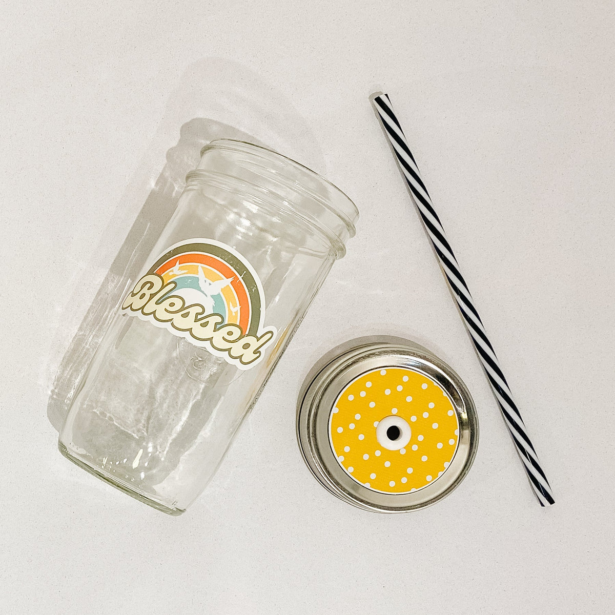 Flat lay of a mason jar tumbler that says "blessed", a yellow straw lid with white polka dots, and a black and white striped straw.