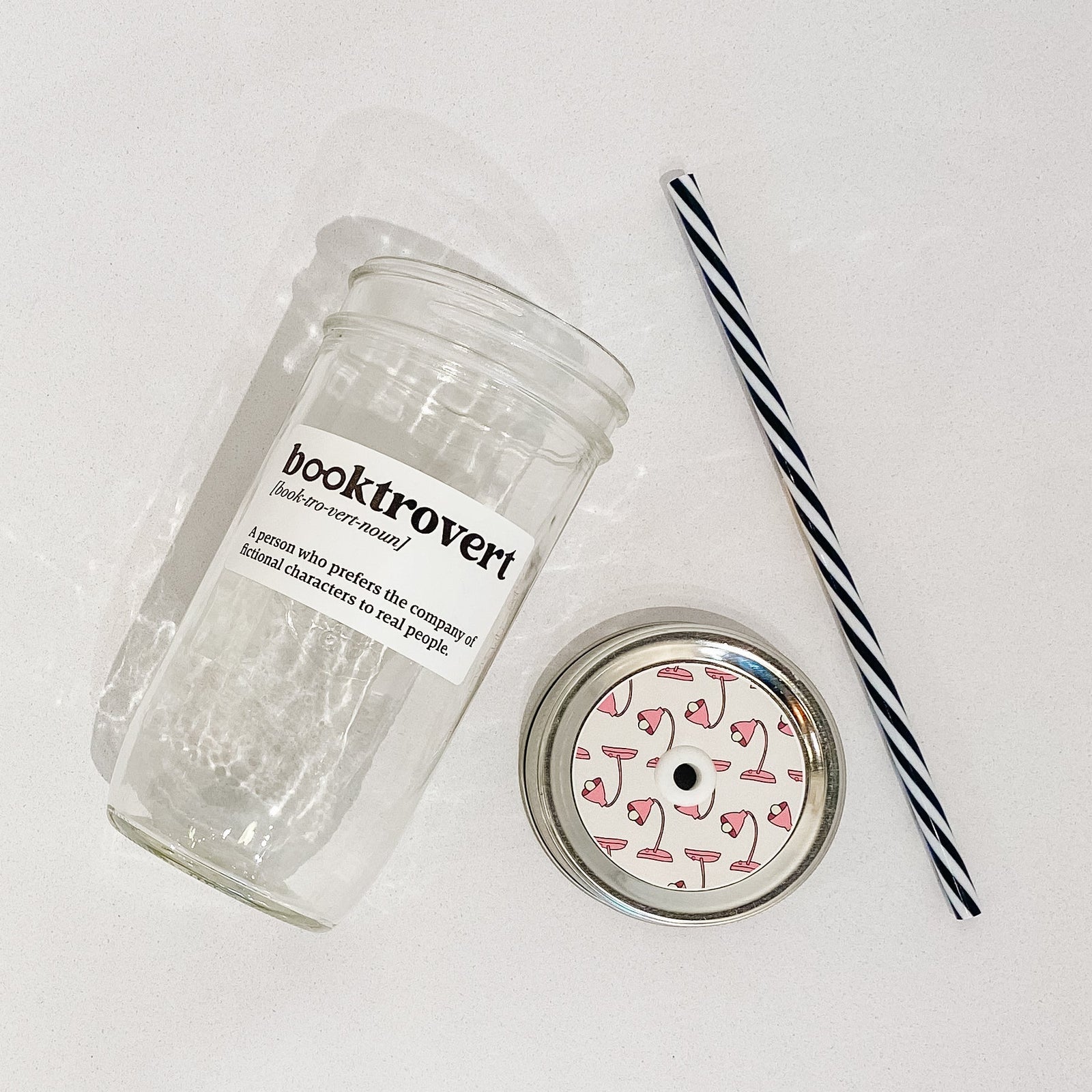 Flat lay of a mason jar tumbler that says “booktrovert. A person who prefers the company of fictional characters to real people”, and straw lid with a repeating pattern of a pink lamp, and a black and white striped straw.
