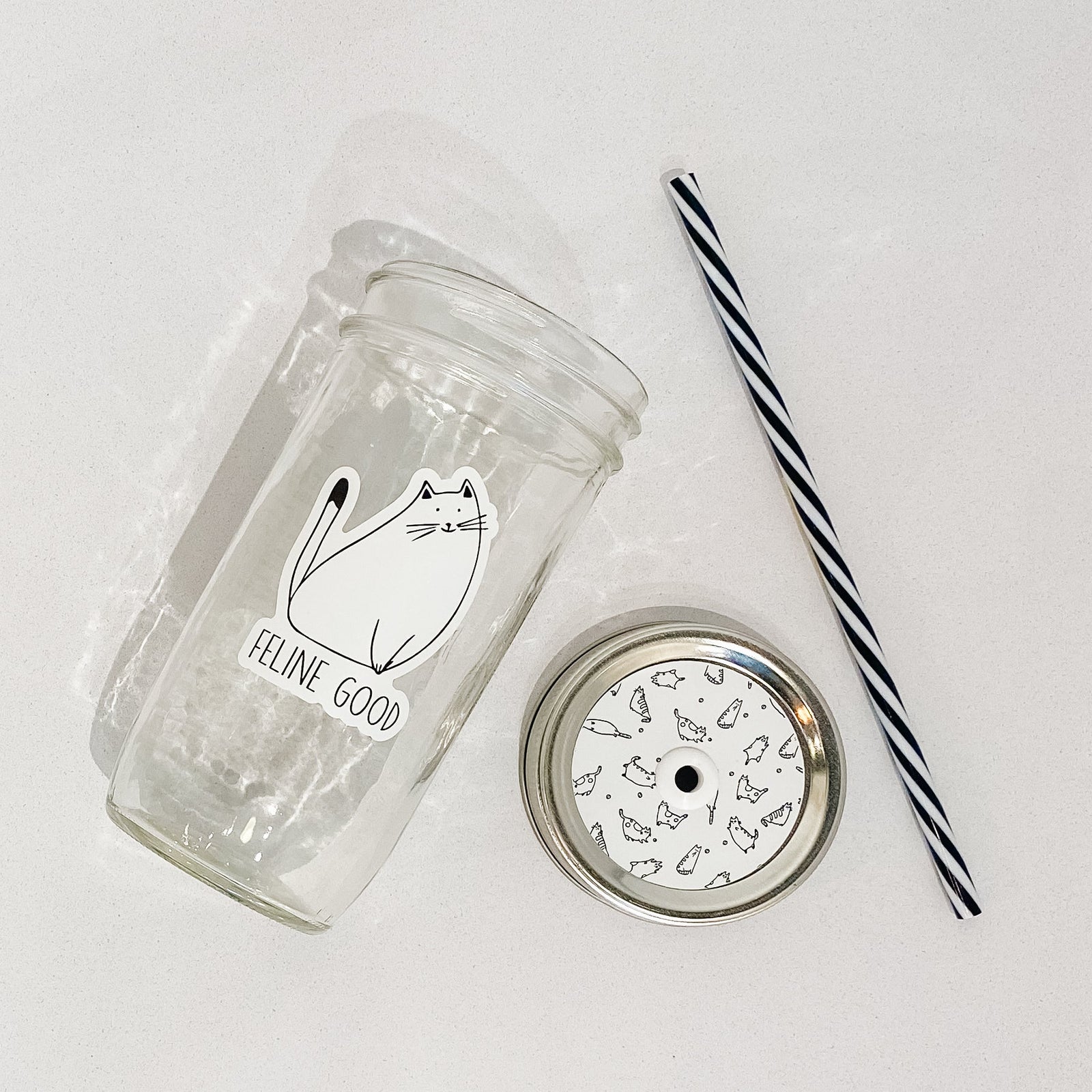 Flat lay of a mason jar tumbler that says “feline good”, a white straw lid with black outlines of cats repeating, and a black and white striped straw.