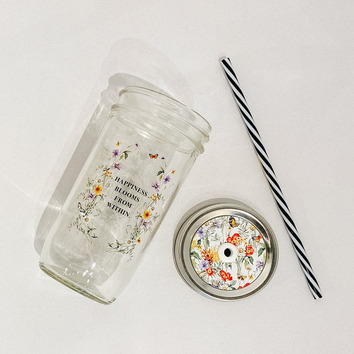 Flat lay of a mason jar tumbler that says “happiness blooms from within”, a straw lid that is busy with different flowers, greenery, and butterflies, and a black and white straw.