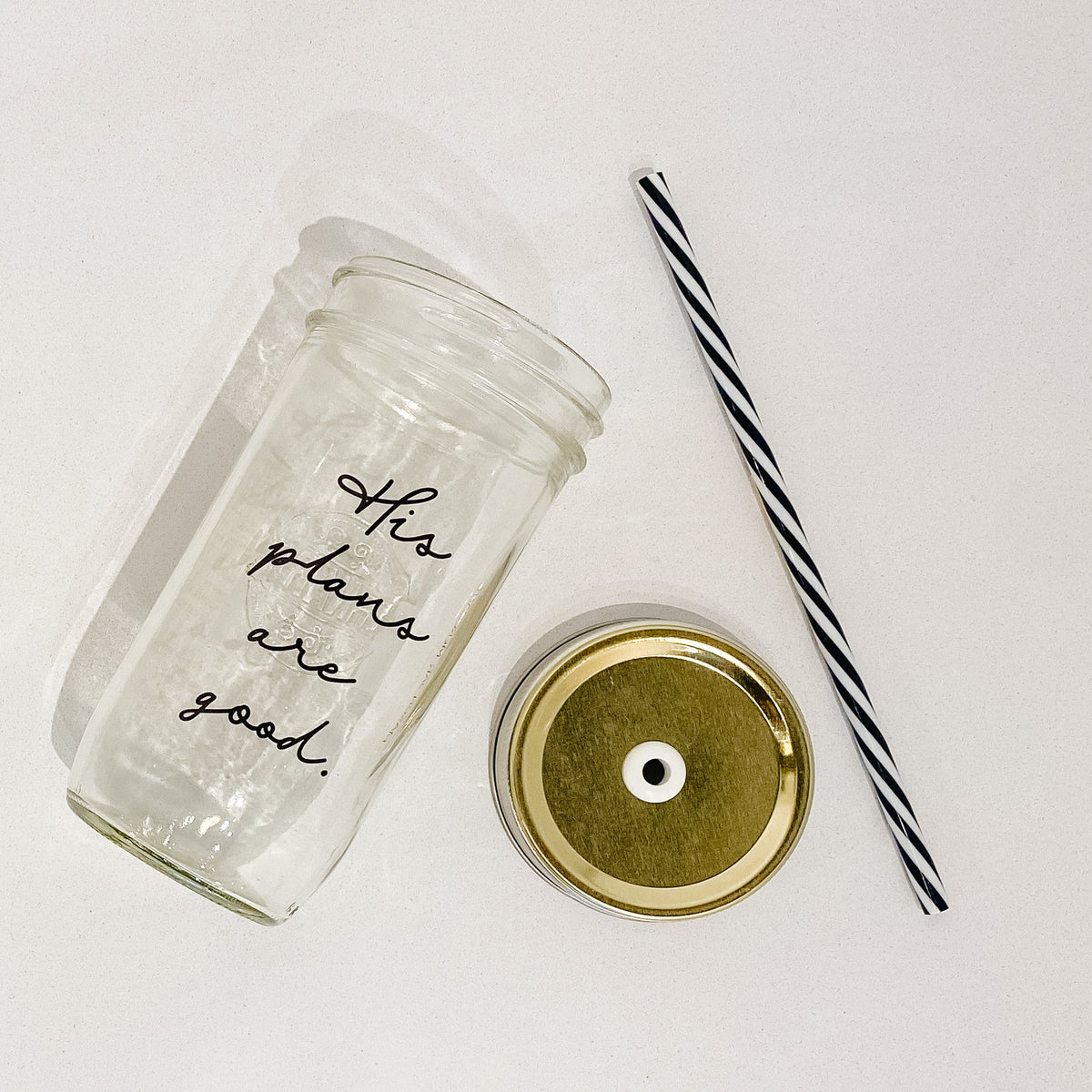 Flat lay of a mason jar tumbler that says “his plans are good”, a gold straw lid, and a black and white striped straw