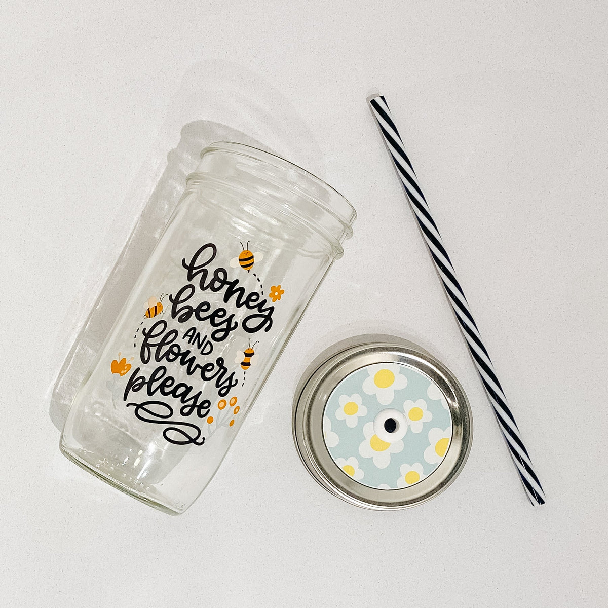 Flat lay of a mason jar tumbler that says “honey, bees, and flowers please”, a straw lid that has a light blue background and white flowers with yellow centres, and a black and white striped straw.