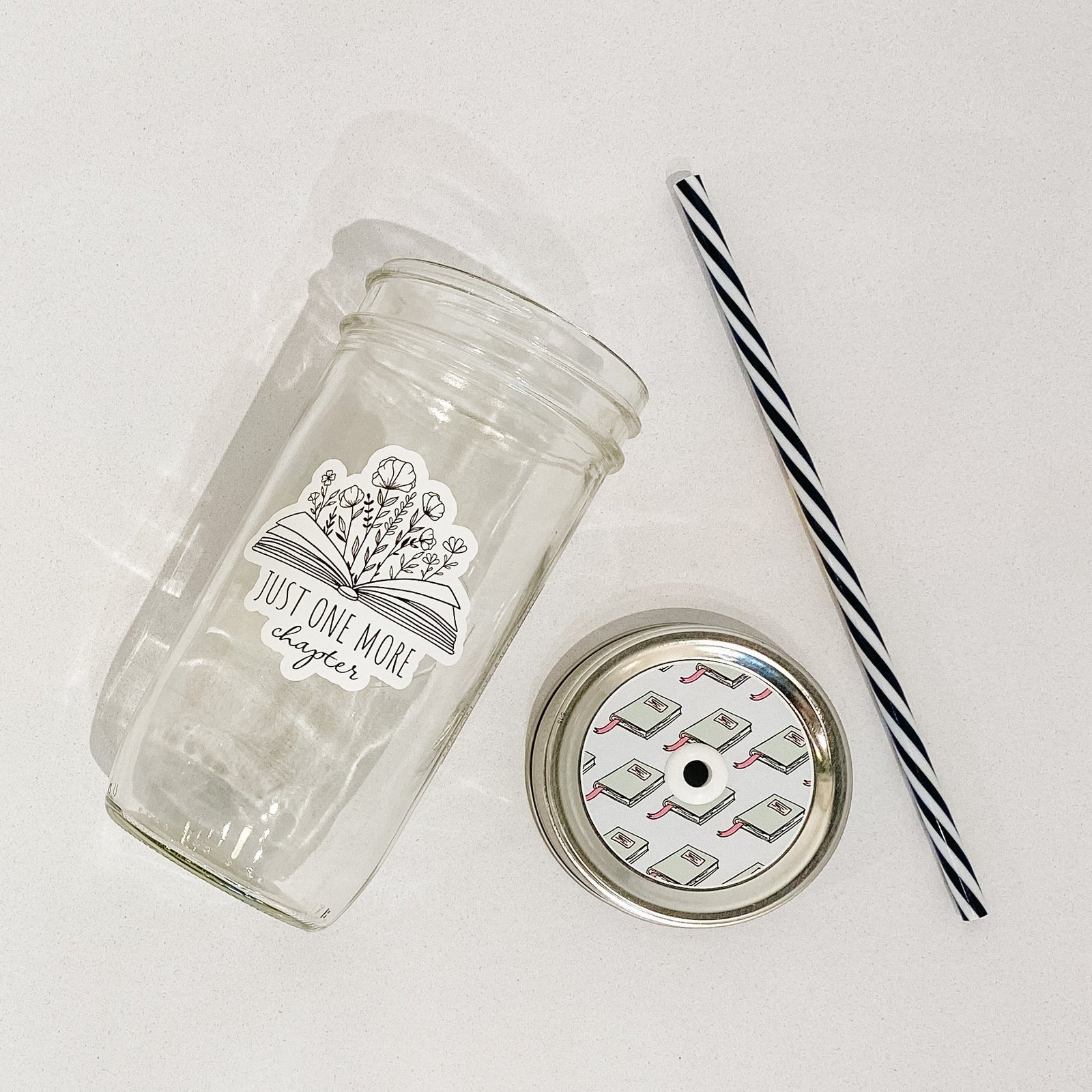 Flat lay of a mason jar tumbler that says “just one more chapter” with flowers growing out of an open book, a straw lid that has a repeating pattern of a green book, and a black and white striped straw.