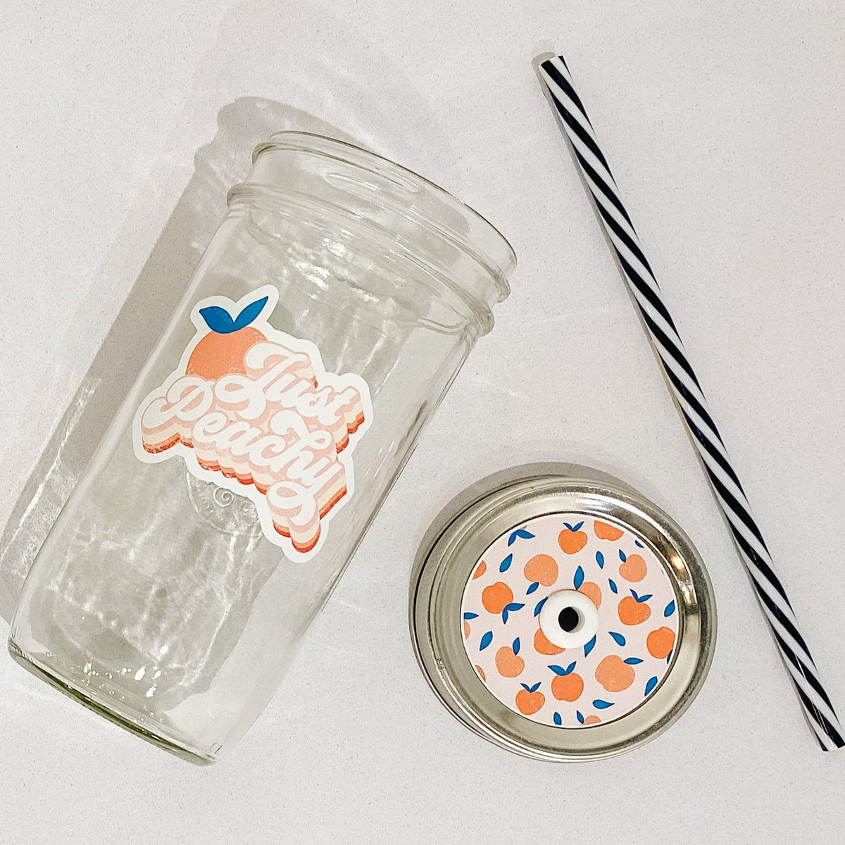 Flat lay of a mason jar tumbler that reads “just peachy”, a straw lid with a repeating peach pattern, and a black and white striped straw.