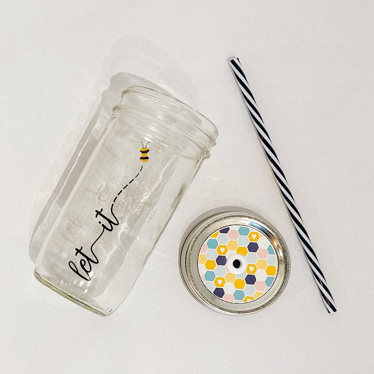 Flat lay of a mason jar tumbler that says “let it bee” with the bee as a drawing, a straw lid with multi colour hexagons to depict a honeycomb, and a black and white striped straw