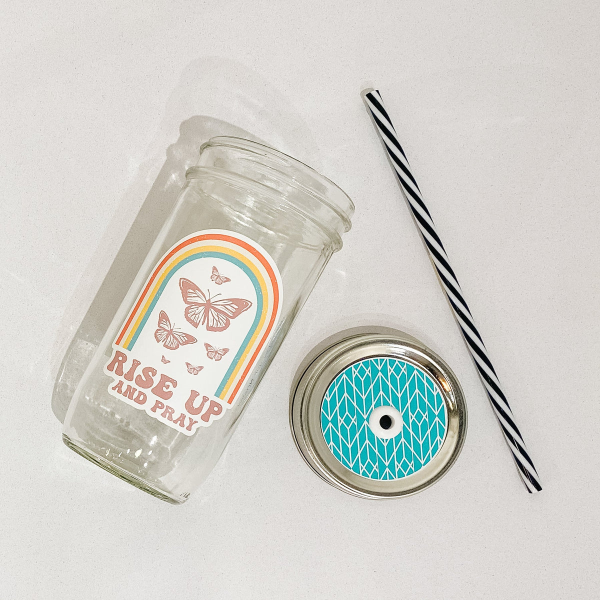 Flat lay of mason jar tumbler that says "Rise up and pray", a blue straw lid with a white chevron-style pattern, and a black and white striped straw.