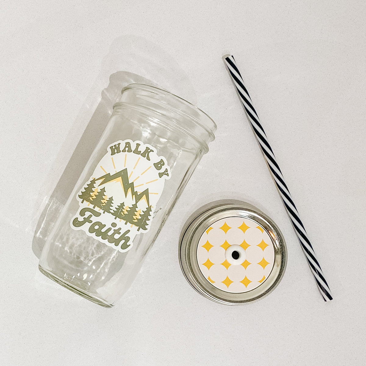Flat lay of a mason jar tumbler that says "walk by faith", a cream colored straw lid with a repeating pattern of star-like diamonds (yellow), and a black and white straw.
