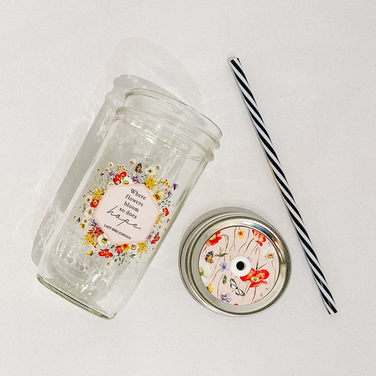 Flat lay of a mason jar tumbler that says “Where flowers bloom so does hope. Lady Bird Johnson”, a pink straw lid with flowers and butterflies, and a black and white striped straw.