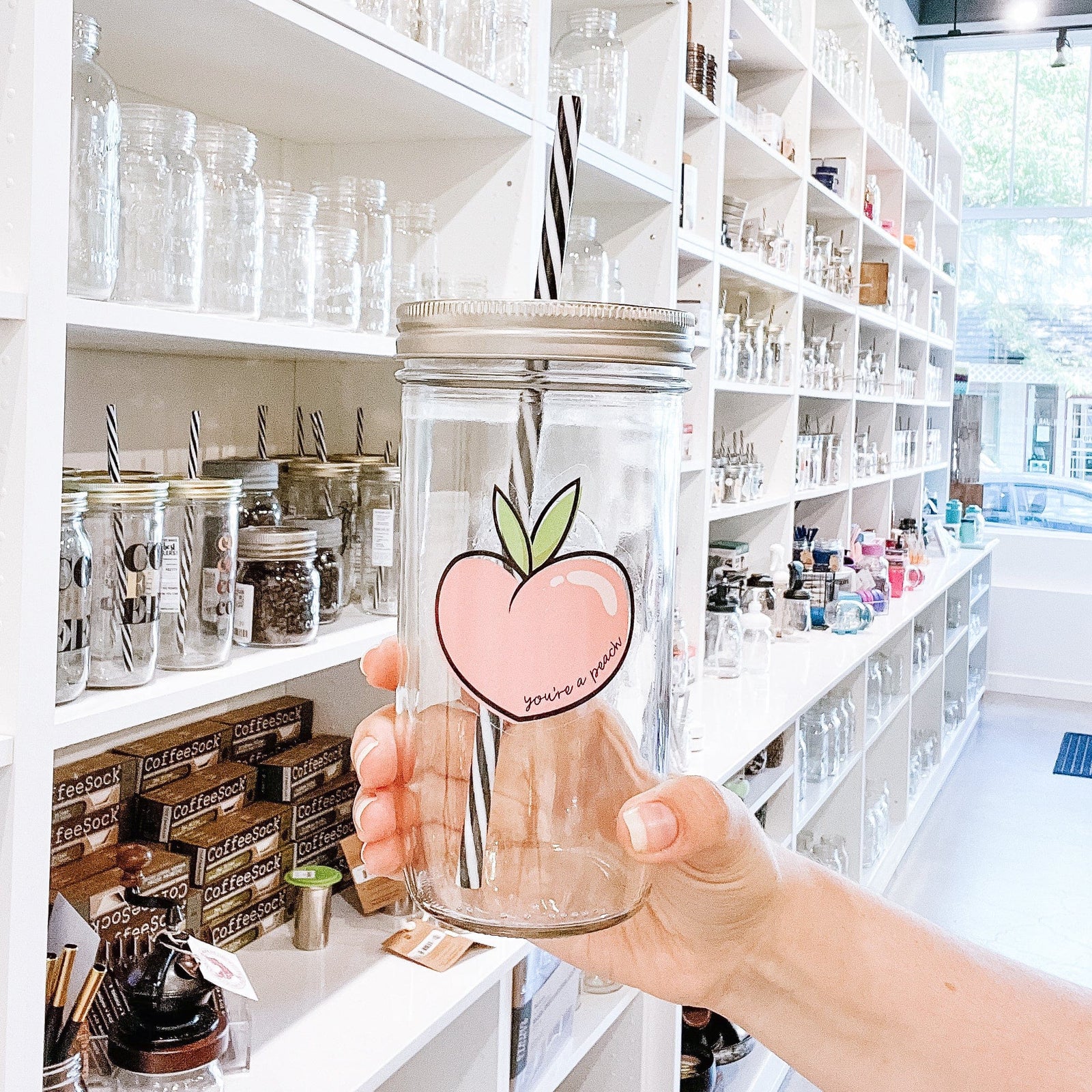 A woman holding a transparent mason jar tumbler with a cute drawing or picture of a peach. Inside the peach is a text that says: You're a peach." It has a black and white straw and behind it is a white cabinet or a storage full of different types of mason jar tumblers.