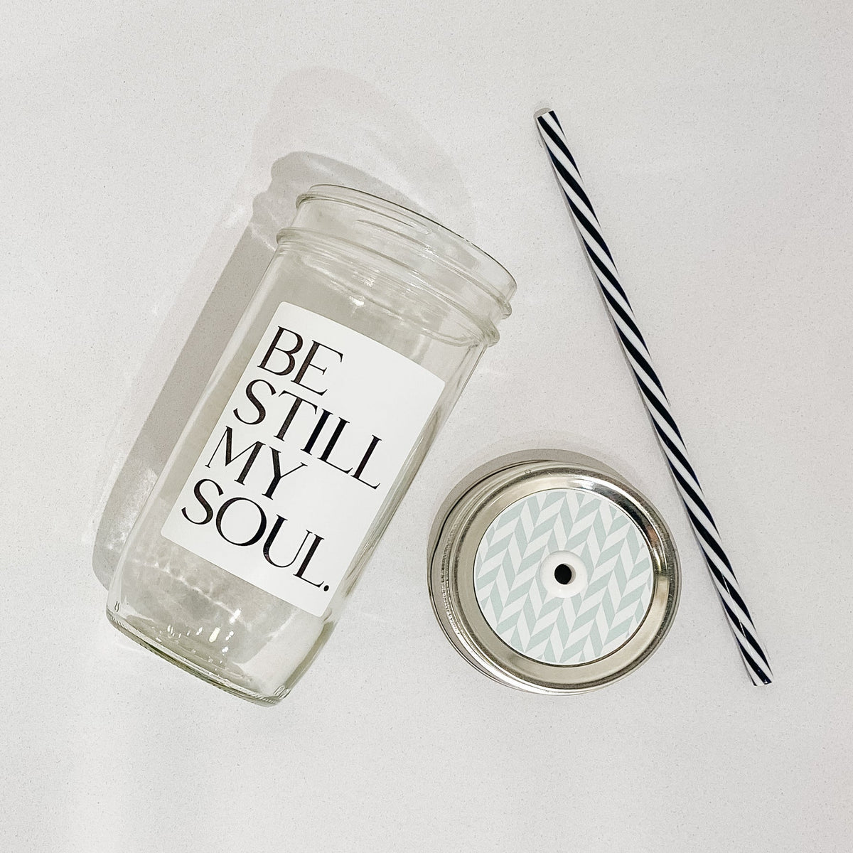 Flat lay of a mason jar tumbler that says "be still my soul”, a straw lid that has a mint and white chevron-esque pattern, and a black and white striped straw.