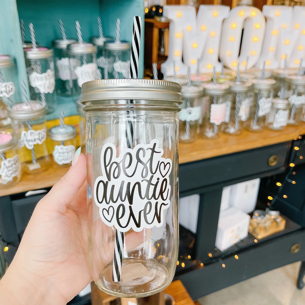 Hand holding a mason jar tumbler that says “best auntie ever” in black script on a white background. There are 2 small hearts beside the words.