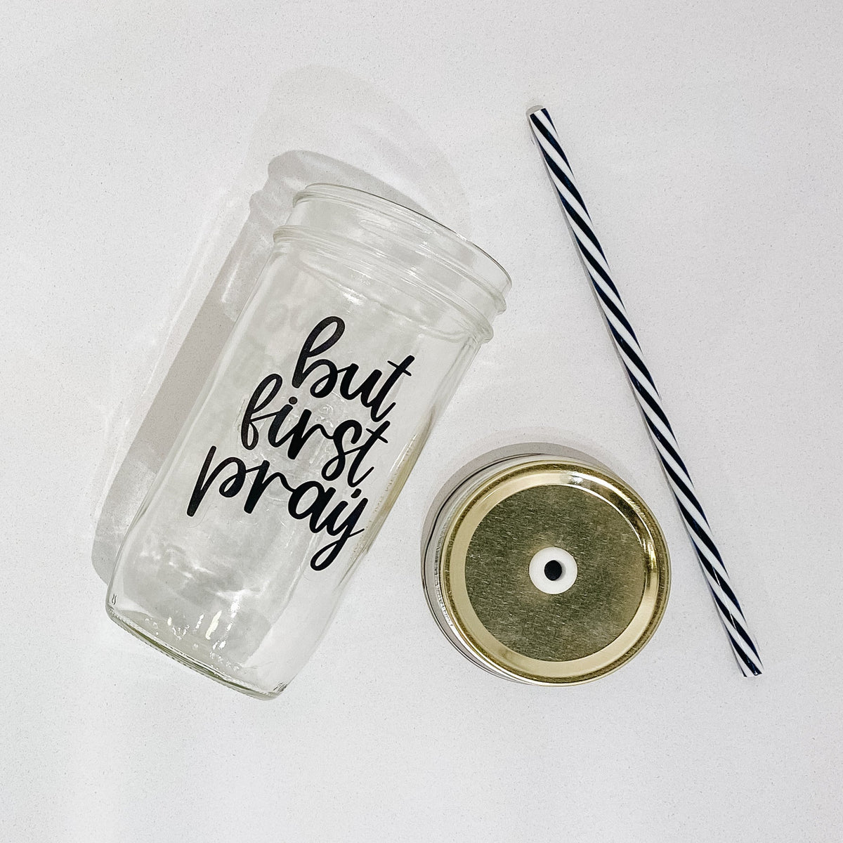 Flat lay of a mason jar tumbler that says “but first, pray”, and straw lid that is gold, and a black and white striped straw.