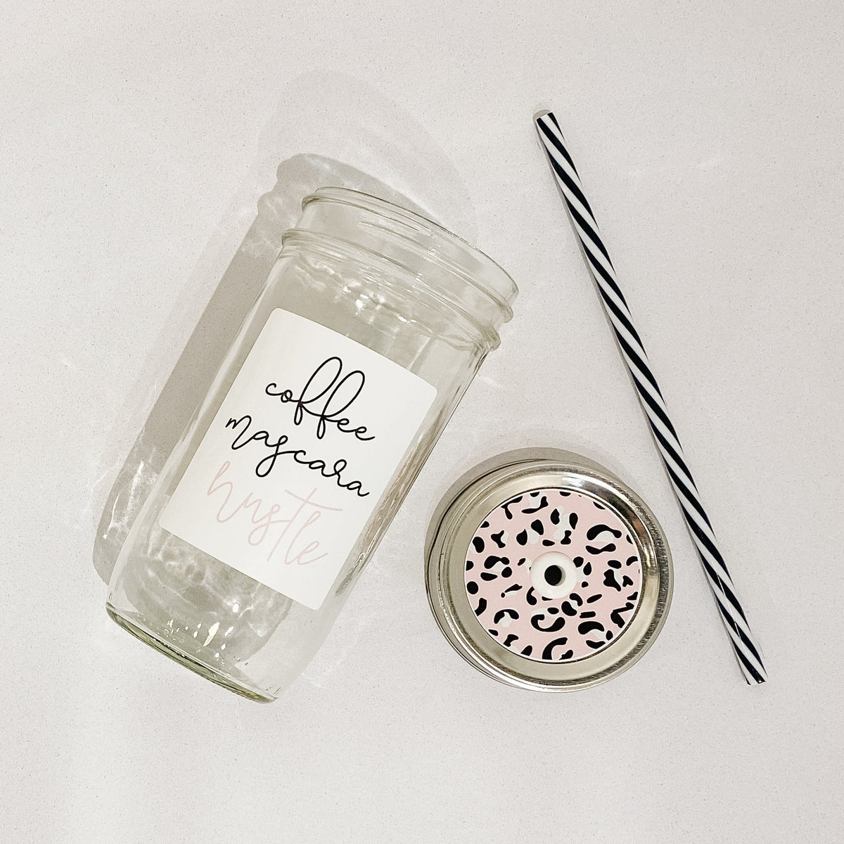Flat lay of a mason jar tumbler that says "coffee mascara hustle”, a straw lid that has a pink cheetah print, and a black and white striped straw.