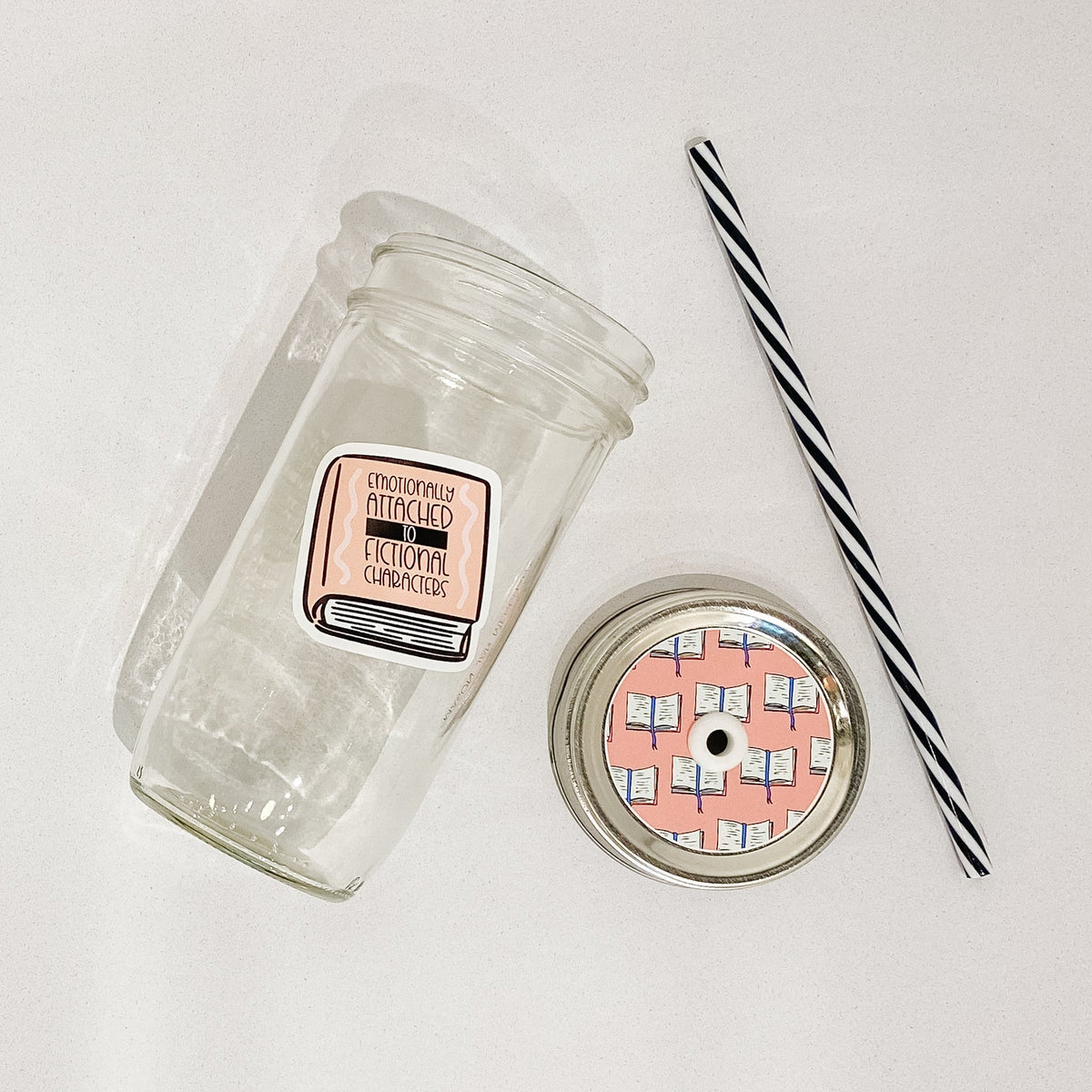 Flat lay of a mason jar tumbler that has an image of a book that says “emotionally attached to fictional character”, a pink straw lid with a repeating pattern of an opened book, and a black and white striped straw.