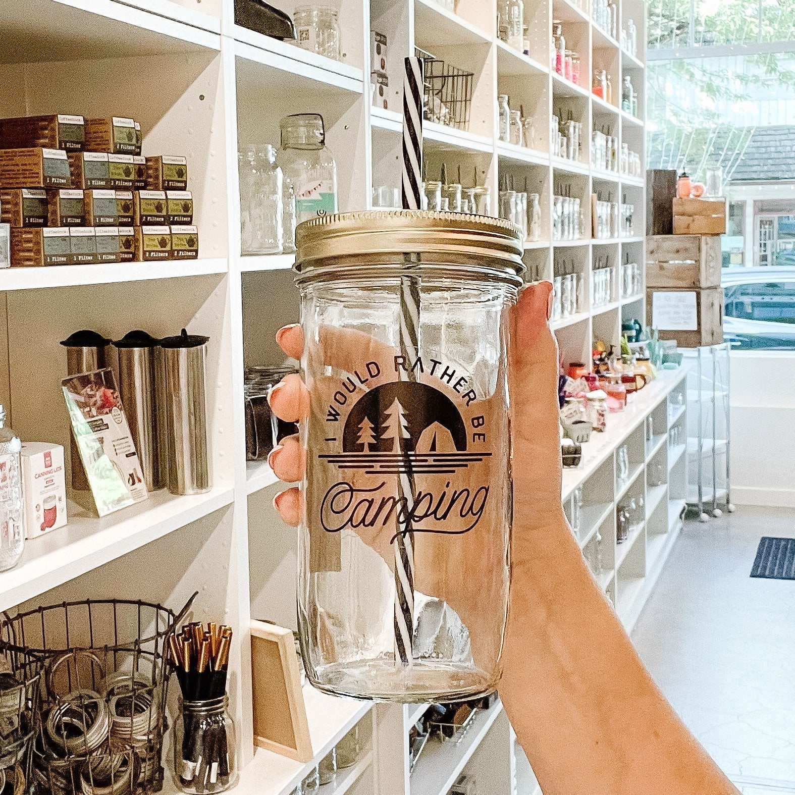 Hand holding a mason jar tumbler that says "I would rather be camping” with clear silhouettes of two trees and a tent on a black background.