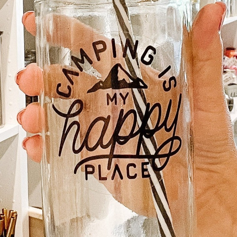 Close up of a hand holding a mason jar tumbler that says “camping is my happy place” with a small mountain peak, all in black on a clear background.