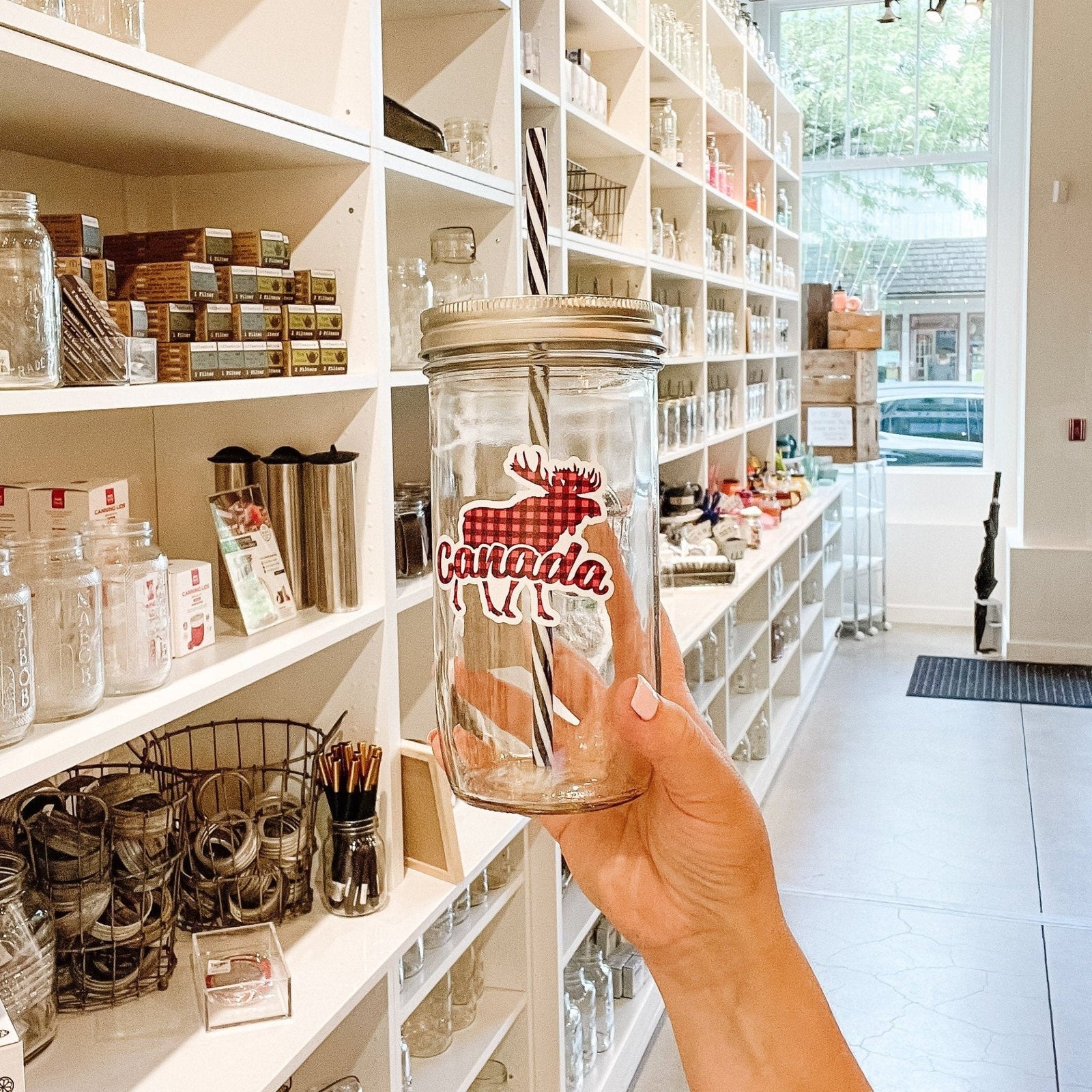 Hand holding a mason jar tumbler that says "Canada" with a silhouette of a moose behind the word. Everything in a red and black plaid pattern.