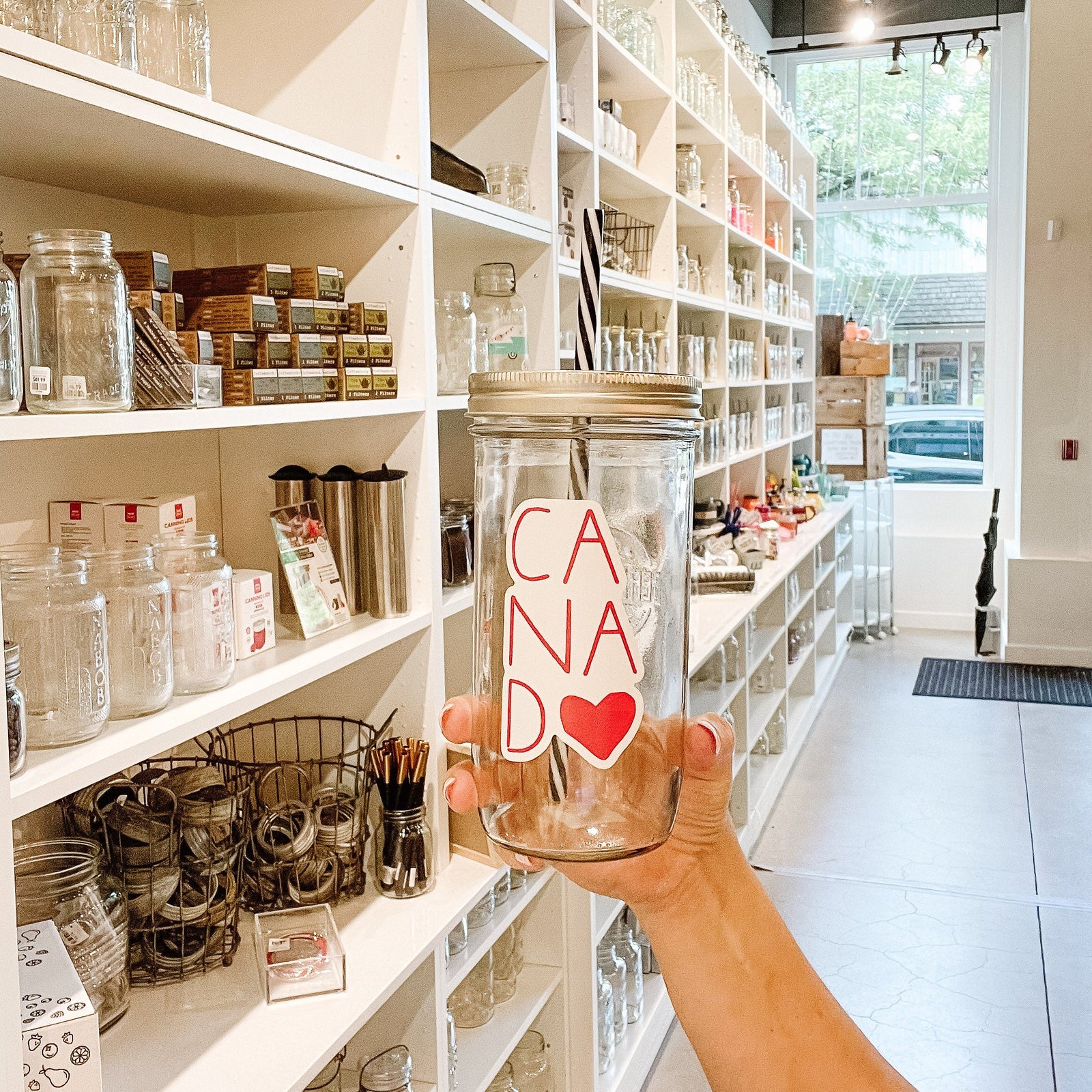 Hand holding a mason jar tumbler that says “canada” with the last A replaced by a heart. All red lettering.