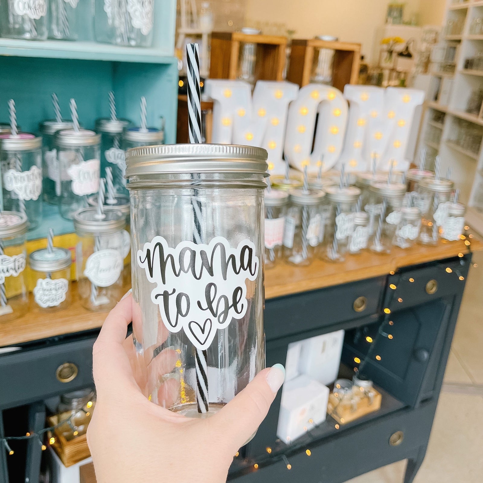 Hand holding a mason jar tumbler that says "mama to be” with a heart under the text. All in black script on a white background