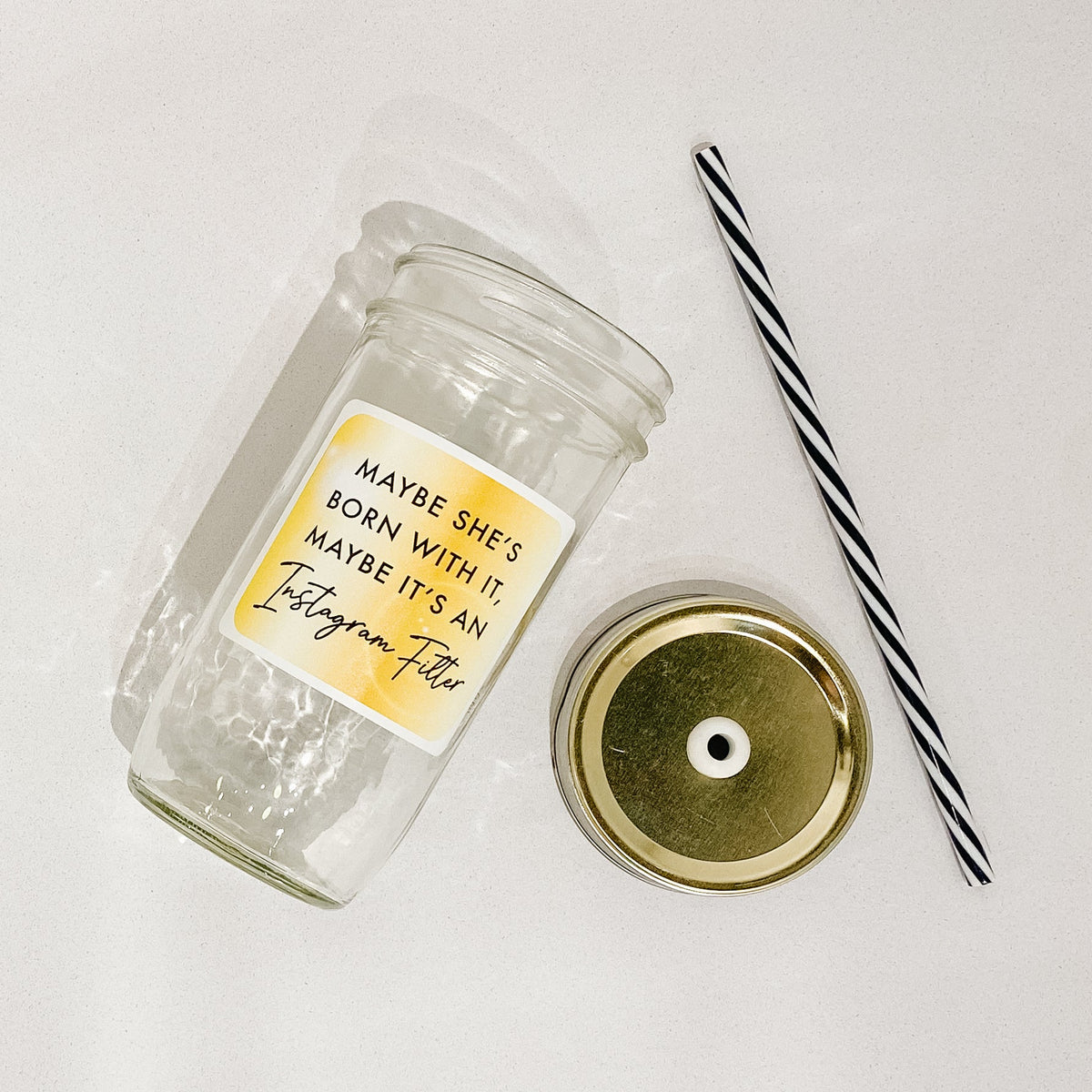 Flat lay of a mason jar tumbler that says “maybe she’s born with in, maybe it’s an instagram filter”, a gold straw lid, and a black and white striped straw