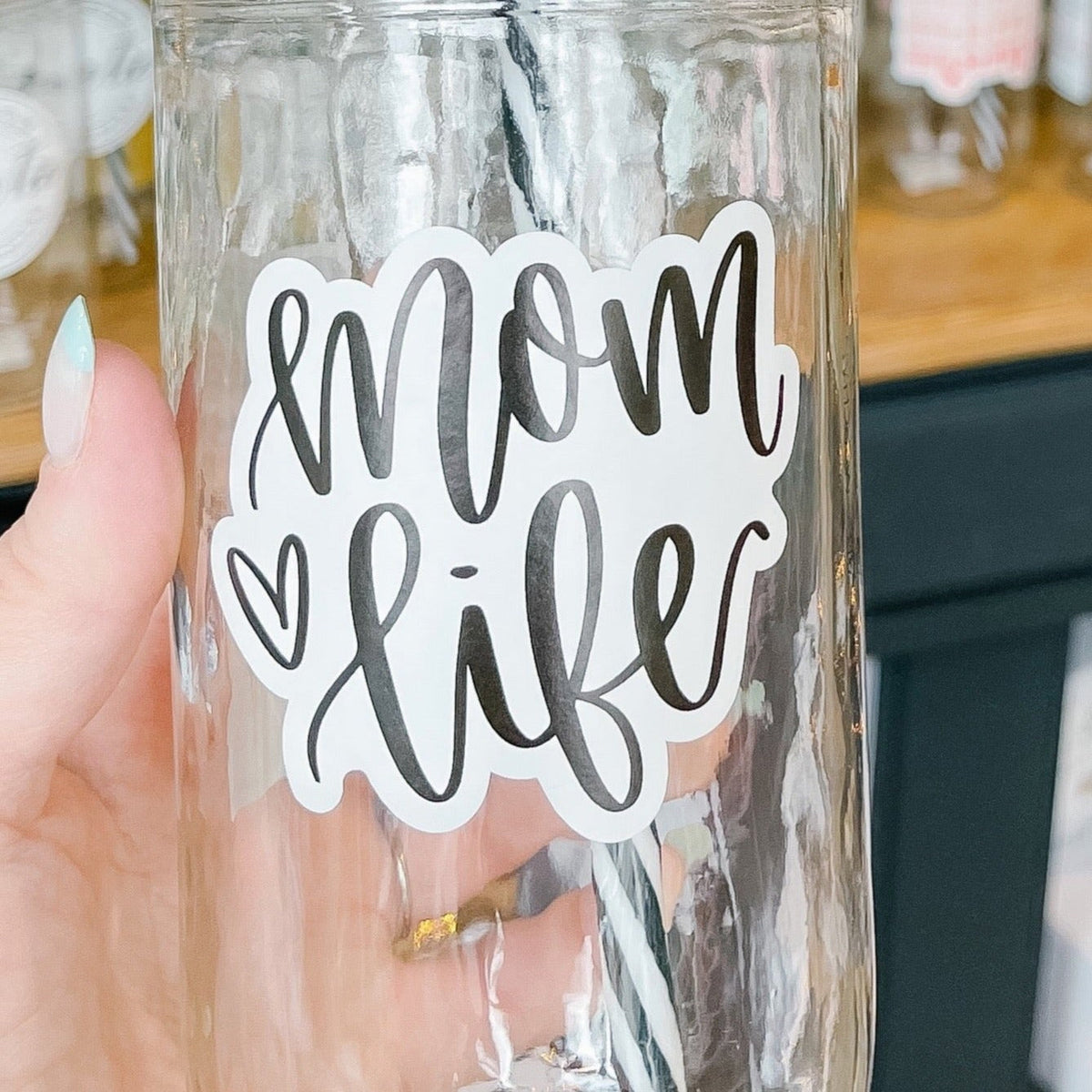 Close up of a hand holding a mason jar tumbler that says “mom life” in black script on a white background, there is a small heart to the left of the text