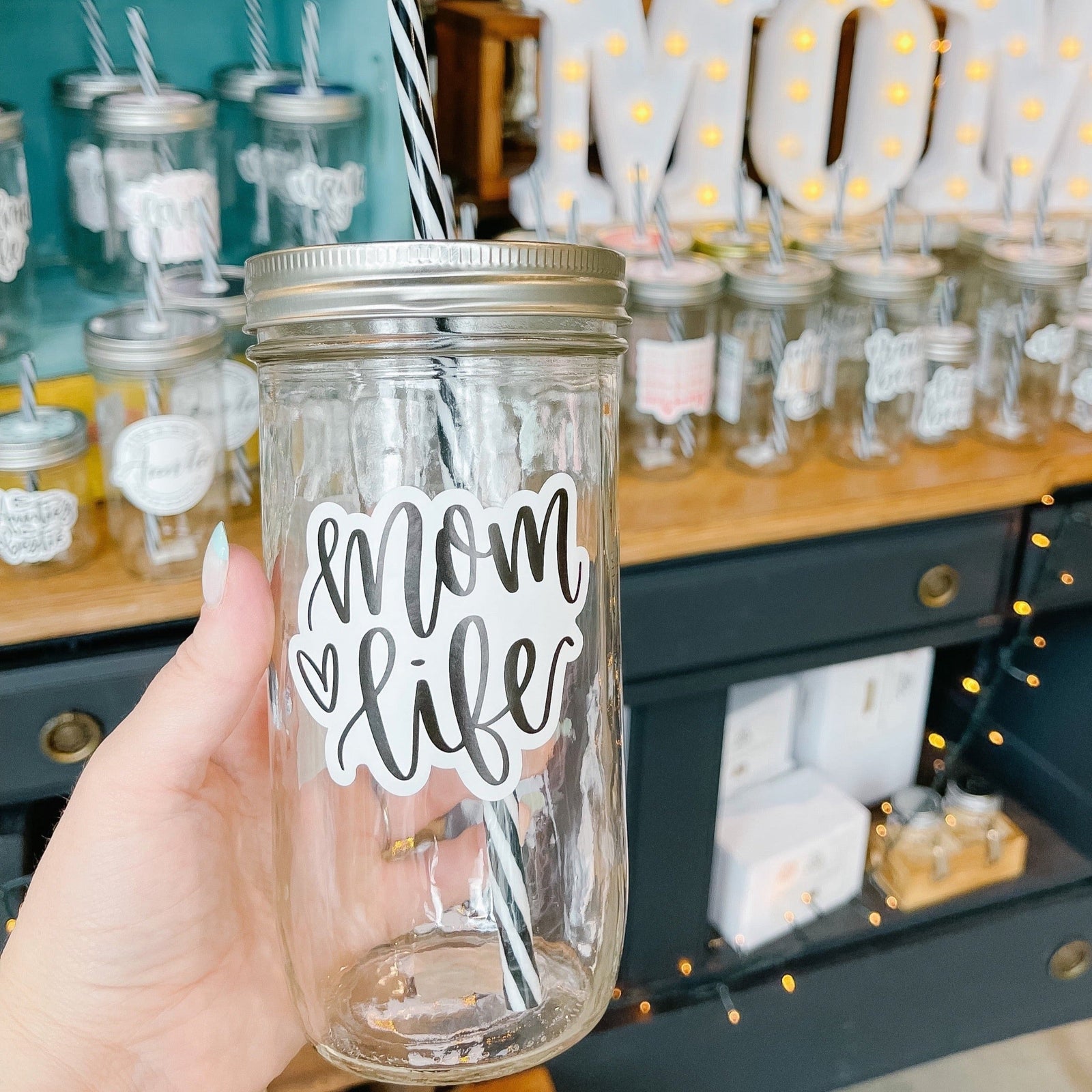 Hand holding a mason jar tumbler that says “mom life” in black script on a white background, there is a small heart to the left of the text