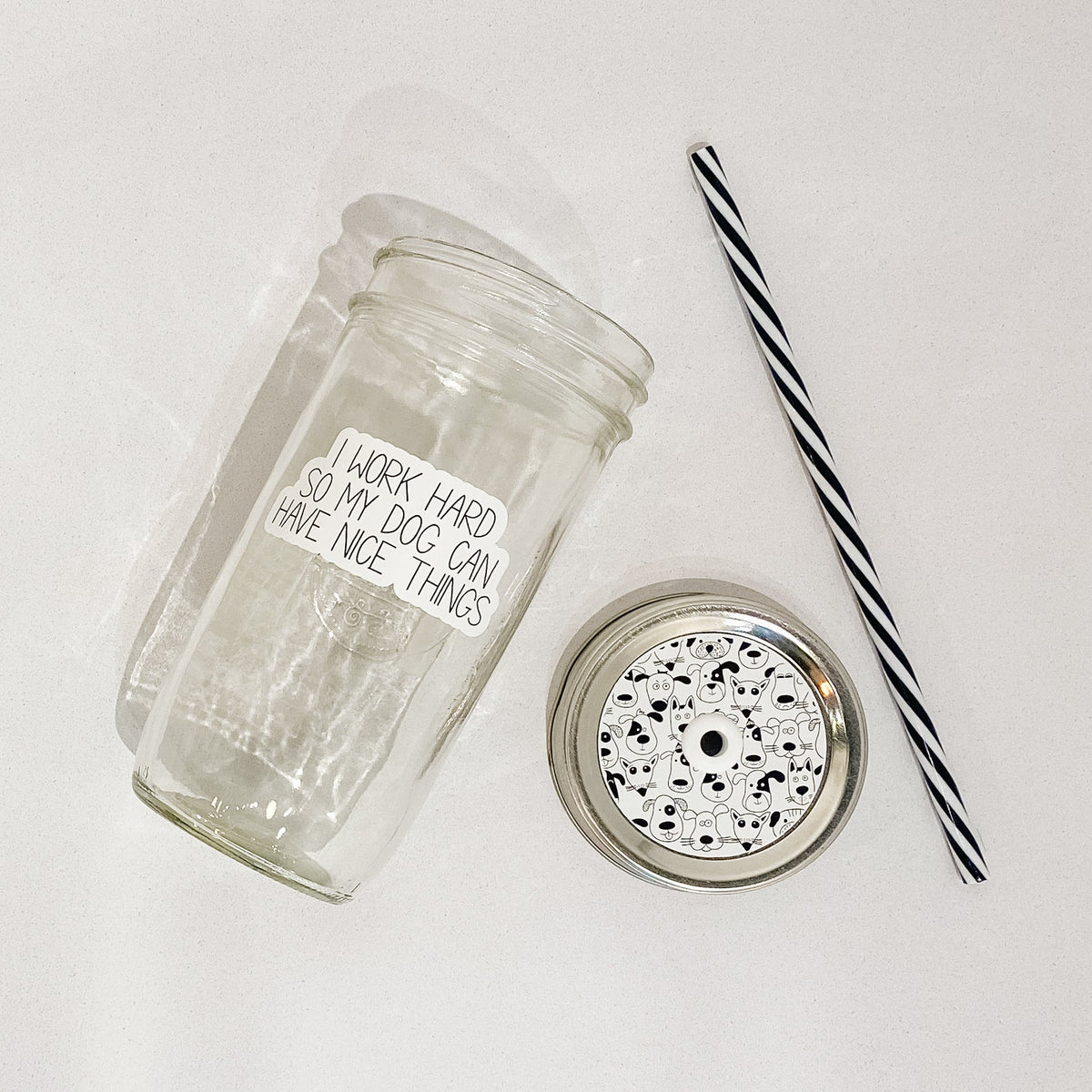 Flat lay of a mason jar tumbler that says “I work hard so my dog can have nice things”, a straw lid that has different dog faces on it, and a black and white striped straw.
