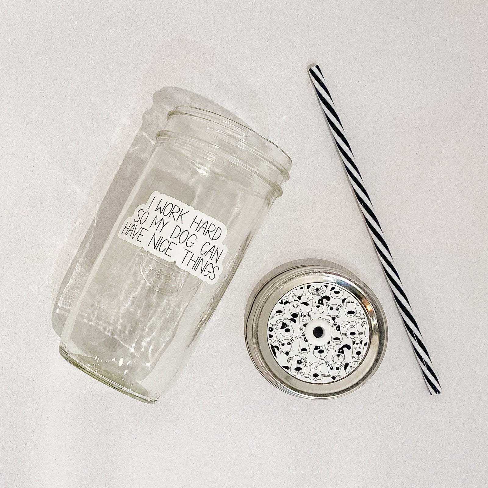 Flat lay of a mason jar tumbler that says “I work hard so my dog can have nice things”, a straw lid that has different dog faces on it, and a black and white striped straw.