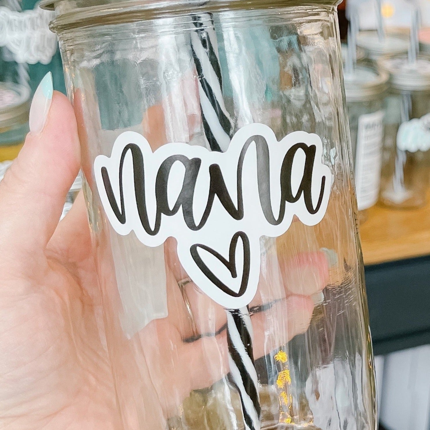Hand holding a mason jar tumbler that says “nana” with a heart under the text. Text in black script on a white background