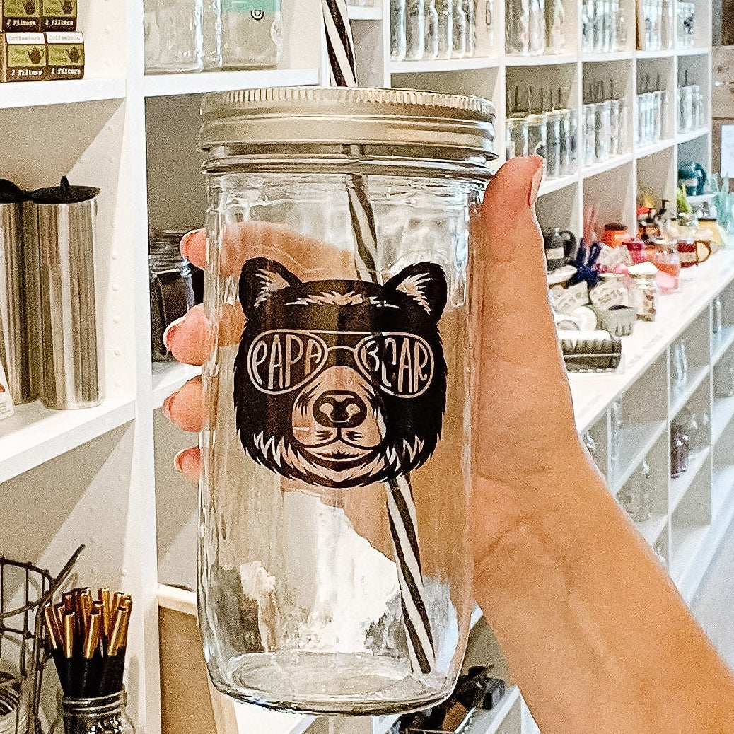 Hand holding a glass tumbler with a bear design and 'Pappy Bear' branding in a store setting.
