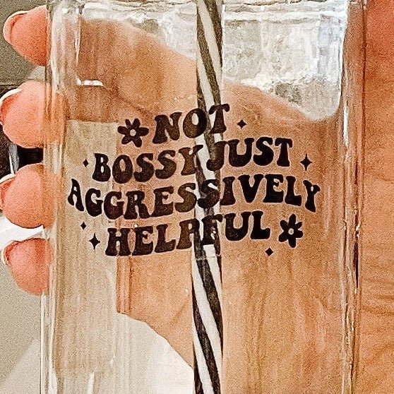 Hand holding a mason jar tumbler that says “not bossy just aggressively helpful” with a small flower at the beginning of the phrase and at the end. All in black.