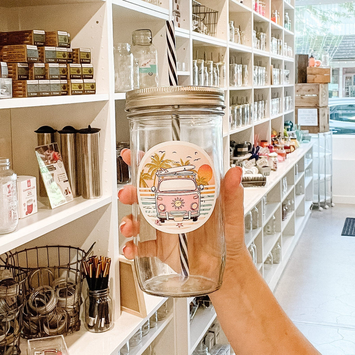 Close up of a hand holding a mason jar tumbler that has a circular decal of an old VW-style bus in front of a sunset with a surfboard on the roof.