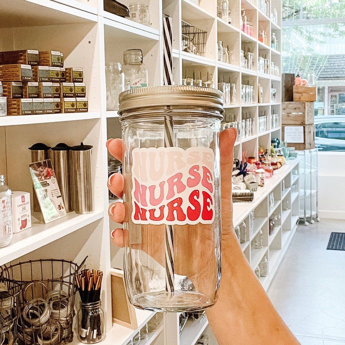 Close up of a hand holding a mason jar tumbler that says “nurse" three times in three different shades of pink.