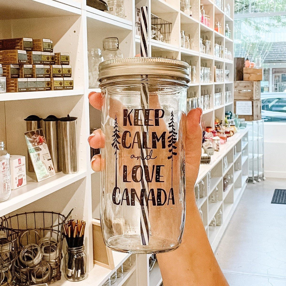 Close up of a hand holding a mason jar tumbler that says “Keep calm and love Canada” with a skinny tree on either side of the words
